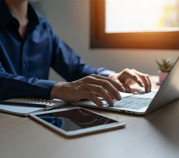 Person typing on a laptop at a desk beside a tablet and a notebook