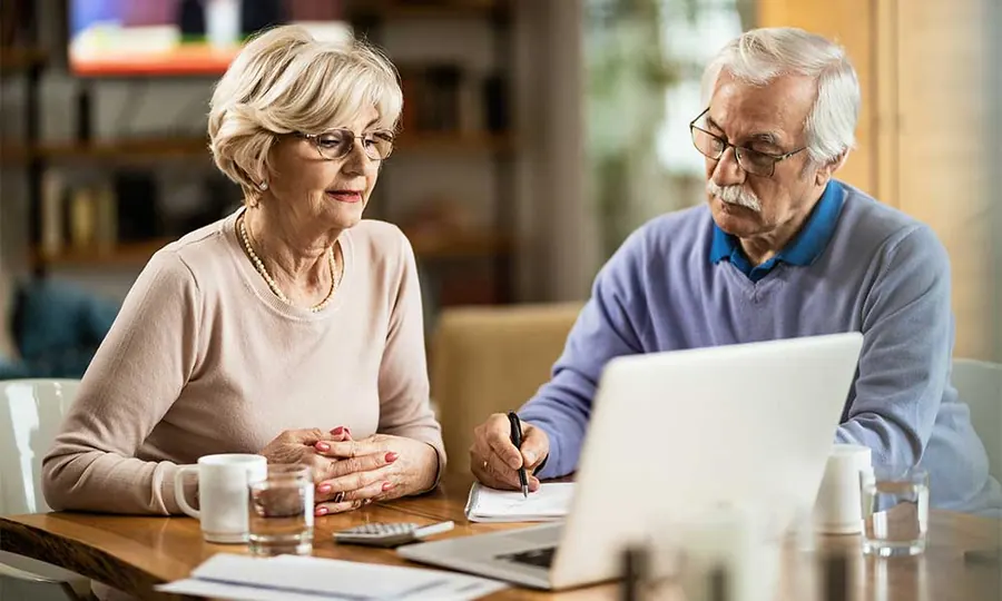Older couple at a dining table reviewing retirement paperwork together beside an open laptop
