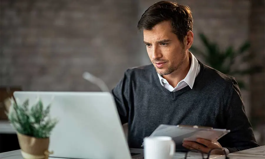 Man at a desk reading a document on a clipboard while looking at his laptop