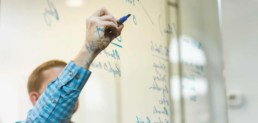 Person in a checked shirt writing notes on a glass whiteboard with a blue marker